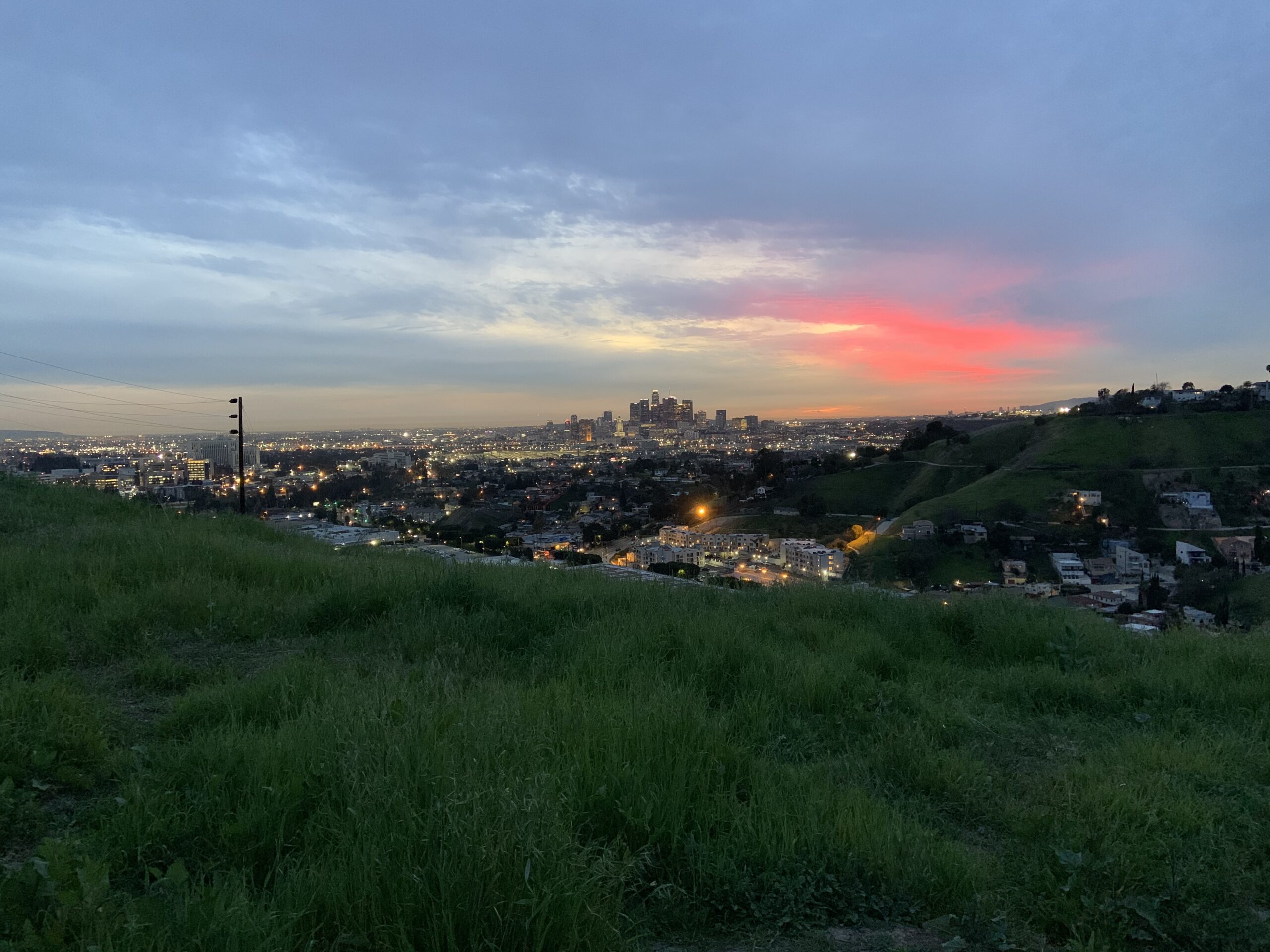 Skyline of Los Angeles with the sun setting in the back, and green hill in the foreground.